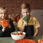 Cute girl and boy with fresh pepper and broccoli in hands standing near desk with assorted vegetables and salad bowl during vegetarian meal preparation