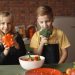 Cute girl and boy with fresh pepper and broccoli in hands standing near desk with assorted vegetables and salad bowl during vegetarian meal preparation